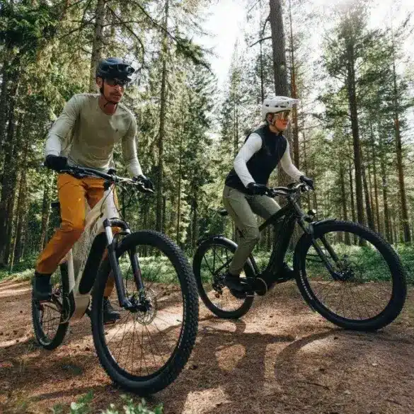 Zwei Personen, ein Mann und eine Frau, fahren nebeneinander auf E-Bikes im Wald auf einem erdigen Weg.