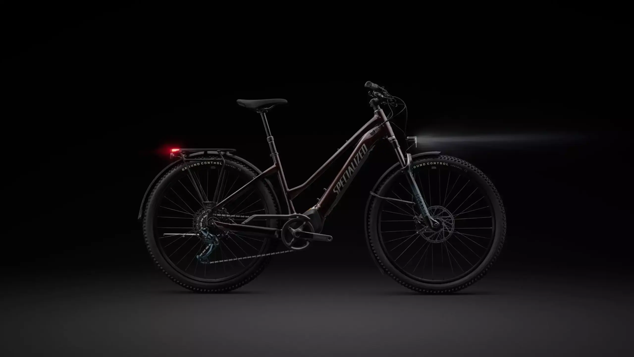 Ein dunkles, mattes E-Bike mit Tiefeinstieg, leuchtendem Front- und Rücklicht, Schutzblechen und Gepäckträger, präsentiert vor schwarzem Hintergrund.