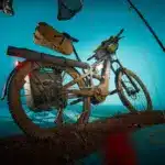 Specialized Turbo Tero X 4.0 E-Bike mit voller Ausstattung und im Schlamm liegend, dargestellt mit einem blauen Hintergrund und umgebenden Gegenständen wie Angelrute und Hut.