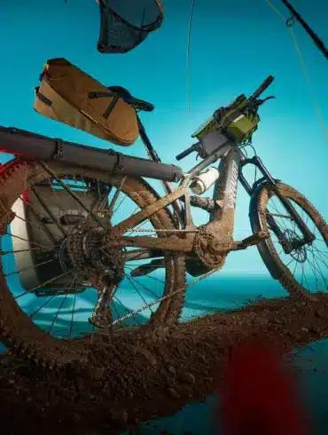 Specialized Turbo Tero X 4.0 E-Bike mit voller Ausstattung und im Schlamm liegend, dargestellt mit einem blauen Hintergrund und umgebenden Gegenständen wie Angelrute und Hut.