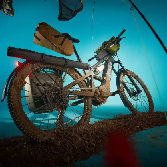 Specialized Turbo Tero X 4.0 E-Bike mit voller Ausstattung und im Schlamm liegend, dargestellt mit einem blauen Hintergrund und umgebenden Gegenständen wie Angelrute und Hut.