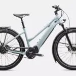 Das Specialized Turbo Vado 4.0 IGH Step-Through ist ein hellgraues E-Bike mit tiefem Einstieg. Es verfügt über Schutzbleche, einen Gepäckträger, eine integrierte Beleuchtung und breite Reifen. Der Rahmen hat eine schlanke Form mit dem Specialized-Logo, und der Akku ist vollständig im Unterrohr integriert. Die Enviolo Nabenschaltung und der Gates Carbon Drive Riemenantrieb sind sichtbar.