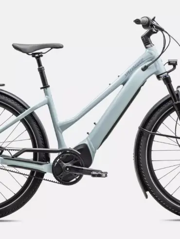 Das Specialized Turbo Vado 4.0 IGH Step-Through ist ein hellgraues E-Bike mit tiefem Einstieg. Es verfügt über Schutzbleche, einen Gepäckträger, eine integrierte Beleuchtung und breite Reifen. Der Rahmen hat eine schlanke Form mit dem Specialized-Logo, und der Akku ist vollständig im Unterrohr integriert. Die Enviolo Nabenschaltung und der Gates Carbon Drive Riemenantrieb sind sichtbar.