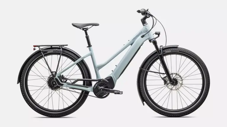 Das Specialized Turbo Vado 4.0 IGH Step-Through ist ein hellgraues E-Bike mit tiefem Einstieg. Es verfügt über Schutzbleche, einen Gepäckträger, eine integrierte Beleuchtung und breite Reifen. Der Rahmen hat eine schlanke Form mit dem Specialized-Logo, und der Akku ist vollständig im Unterrohr integriert. Die Enviolo Nabenschaltung und der Gates Carbon Drive Riemenantrieb sind sichtbar.