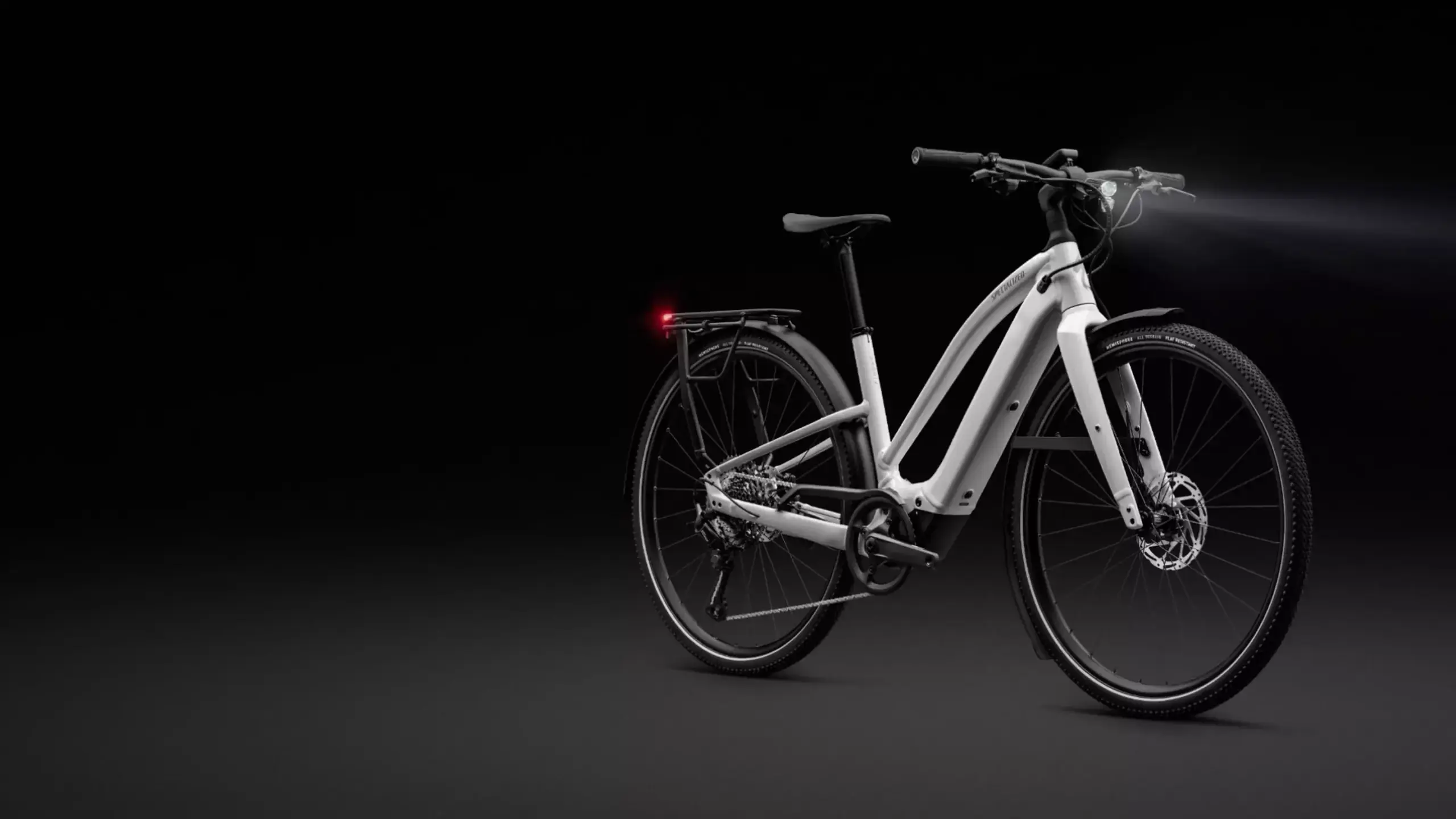 Weißes Specialized Turbo Vado SL 2 5.0 Step-Through EQ E-Bike mit integriertem Akku und Gepäckträger, frontal leicht schräg von vorne mit beleuchtetem Scheinwerfer auf dunklem Hintergrund.