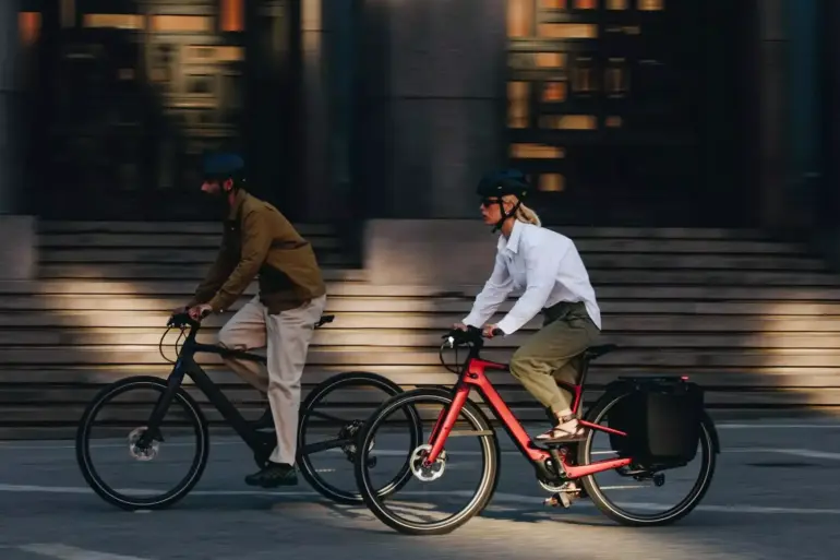 Zwei Personen, eine Frau und ein Mann, fahren E-Bikes nebeneinander auf einer Straße.