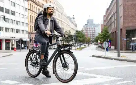 Mann mit Helm und Rucksack fährt ein dunkles Stevens Amant 7.2 HT City-Bike mit Front- und Heckgepäckträger in der Stadt.