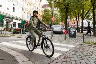 Frau mit Helm und Sonnenbrille fährt ein beiges STEVENS Aviolo 5.2 LT Tourenrad mit Gepäckträger über eine Zebrastreifen-Kreuzung.