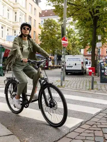 Frau mit Helm und Sonnenbrille fährt ein beiges STEVENS Aviolo 5.2 LT Tourenrad mit Gepäckträger über eine Zebrastreifen-Kreuzung.