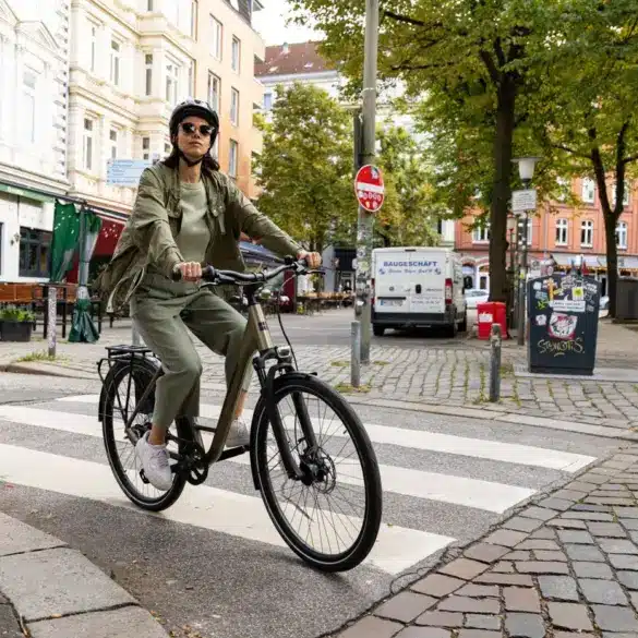 Frau mit Helm und Sonnenbrille fährt ein beiges STEVENS Aviolo 5.2 LT Tourenrad mit Gepäckträger über eine Zebrastreifen-Kreuzung.