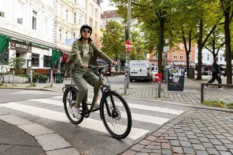 Frau mit Helm und Sonnenbrille fährt ein beiges STEVENS Aviolo 5.2 LT Tourenrad mit Gepäckträger über eine Zebrastreifen-Kreuzung.