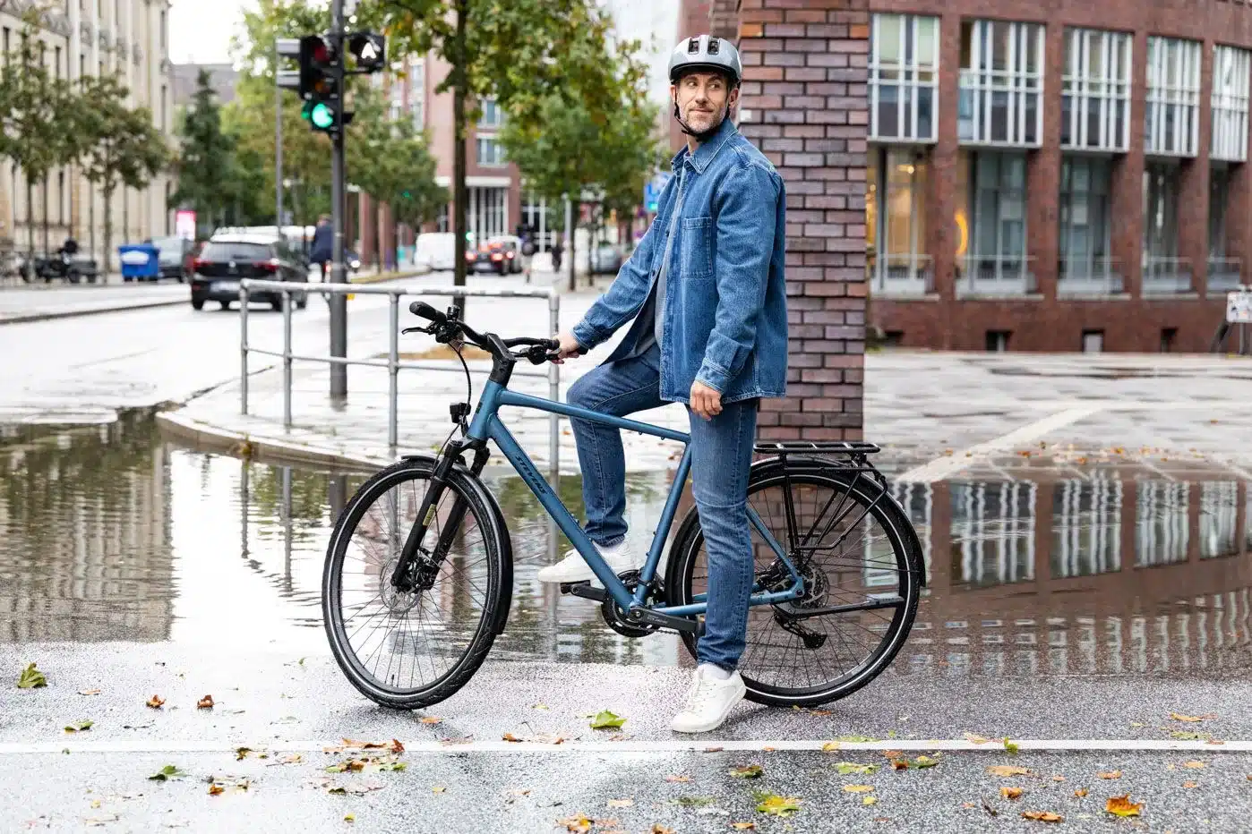Mann auf einem blauen STEVENS Aviolo 8.1 LT Trekkingrad stehend neben einer Ziegelwand mit Spiegelungen auf der nassen Straße im Hintergrund.