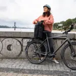 Frau mit Helm und pinkfarbenem Pullover lehnt sich an ihr schwarzes Stevens Caleta 9.1 LT Trekkingrad mit einer schwarzen Gepäcktasche auf dem Gepäckträger.