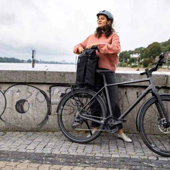 Frau mit Helm und pinkfarbenem Pullover lehnt sich an ihr schwarzes Stevens Caleta 9.1 LT Trekkingrad mit einer schwarzen Gepäcktasche auf dem Gepäckträger.