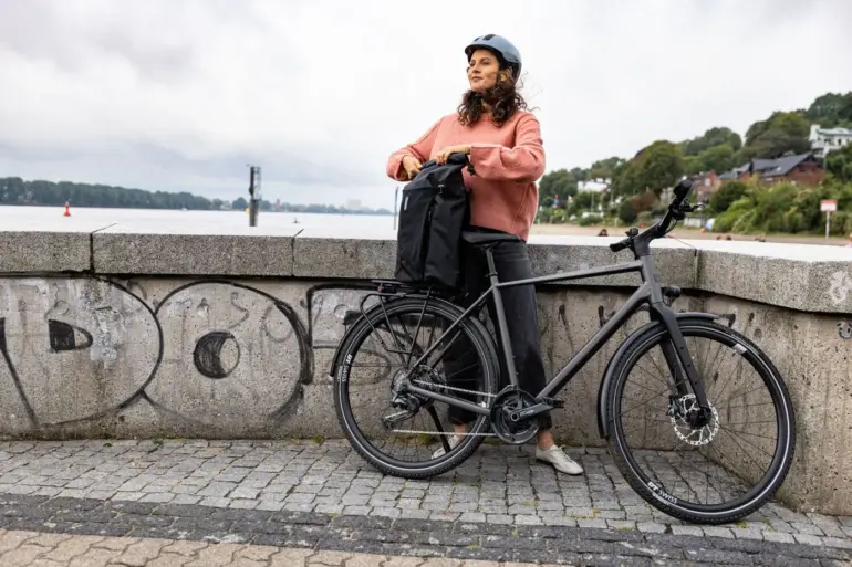 Frau mit Helm und pinkfarbenem Pullover lehnt sich an ihr schwarzes Stevens Caleta 9.1 LT Trekkingrad mit einer schwarzen Gepäcktasche auf dem Gepäckträger.