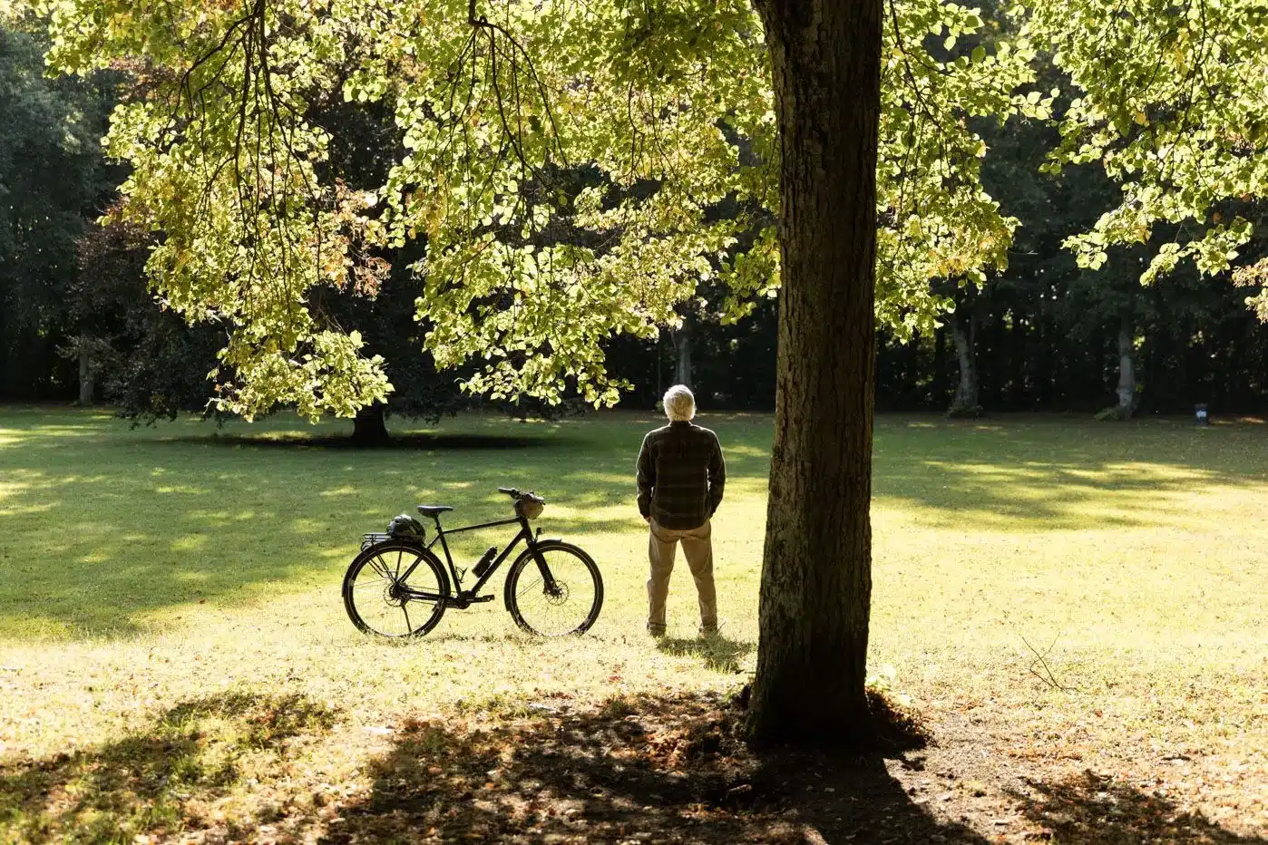 Ein Mann mit einem karierten Hemd und beigen Hosen steht mit dem Rücken zum Betrachter in einem sonnigen Park neben seinem schwarzen Trekkingrad.