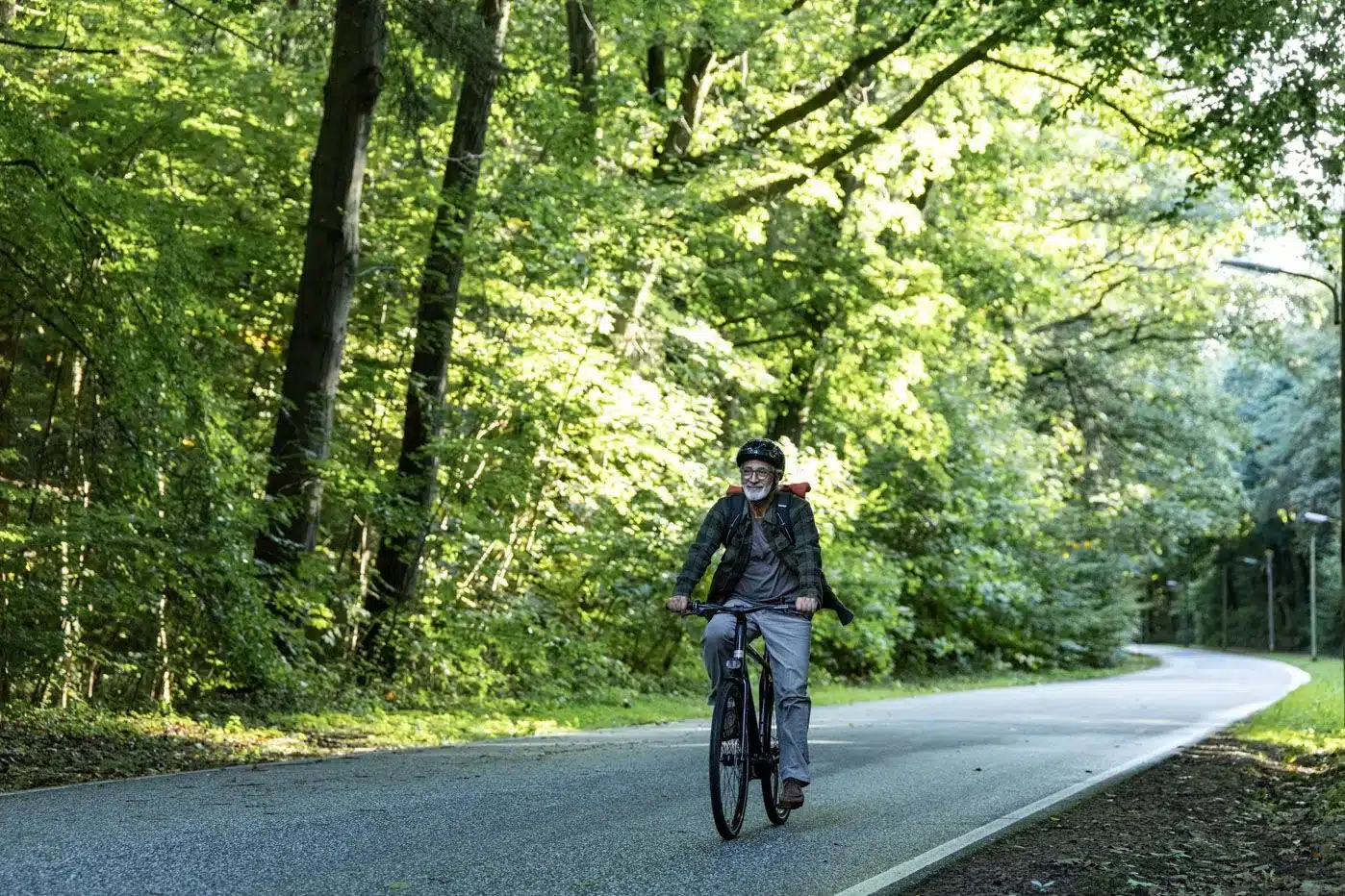 Ein älterer Mann mit Helm und Rucksack fährt auf einem Fahrrad auf einer asphaltierten Straße durch einen beleuchteten Wald.