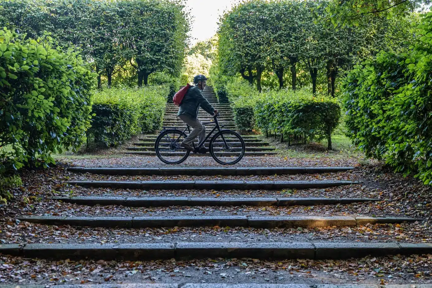 Ein Mann in einem grünen Hemd und mit einem roten Rucksack fährt mit seinem Fahrrad auf einer Steintreppe, gesäumt von Büschen und Bäumen.