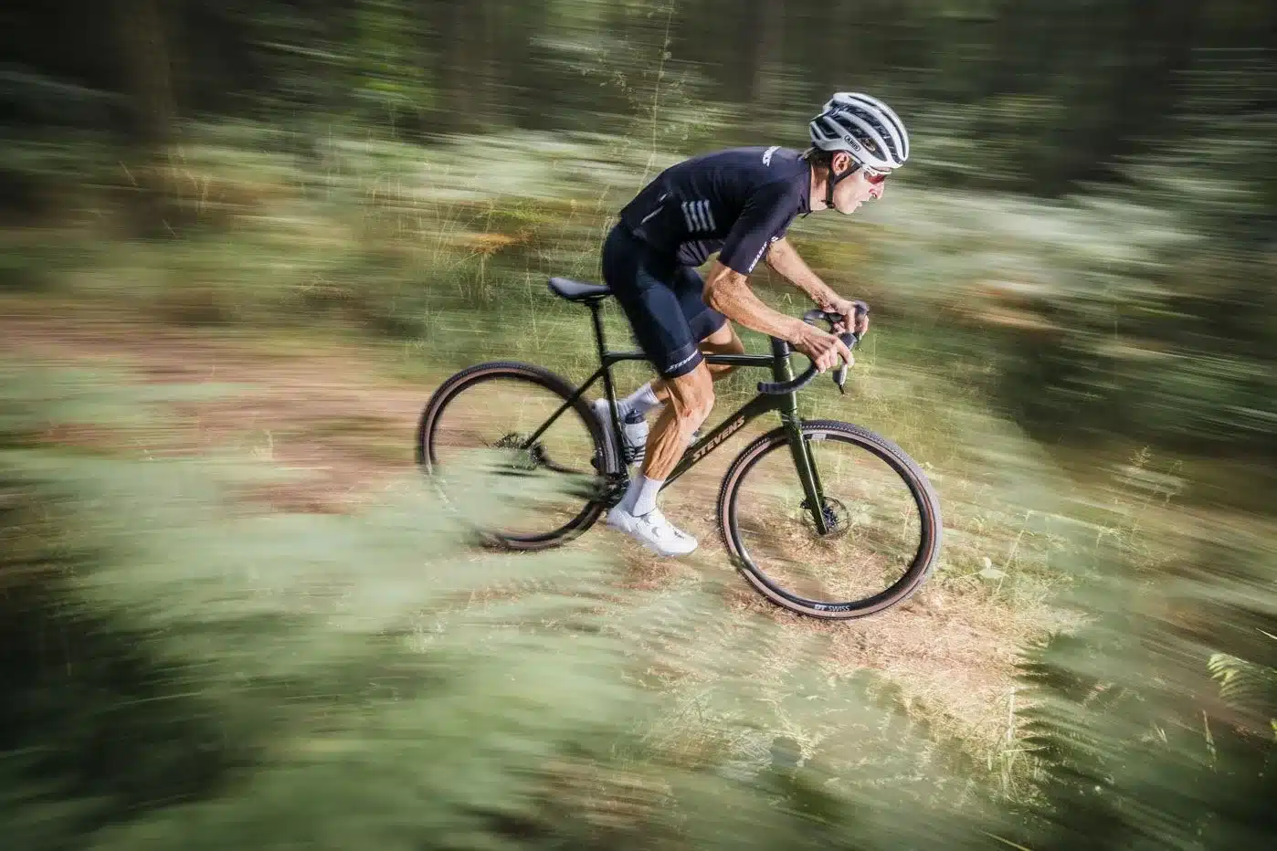 Ein Mann auf einem Stevens Camino RS Gravelbike fährt schnell über einen schlammigen oder erdigen Waldweg, umgeben von verschwommenen grünen Pflanzen und Bäumen. Der Fahrer trägt einen Helm und Cycling-Bekleidung. Die Bewegungsunschärfe erzeugt einen dynamischen Eindruck.