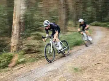 Zwei Radfahrer auf einem Stevens Camino RS Pro Di2 Gravelbike fahren auf einem schmalen Schotterweg durch einen Wald, die Bewegung ist durch eine leichte Bewegungsunschärfe dargestellt.