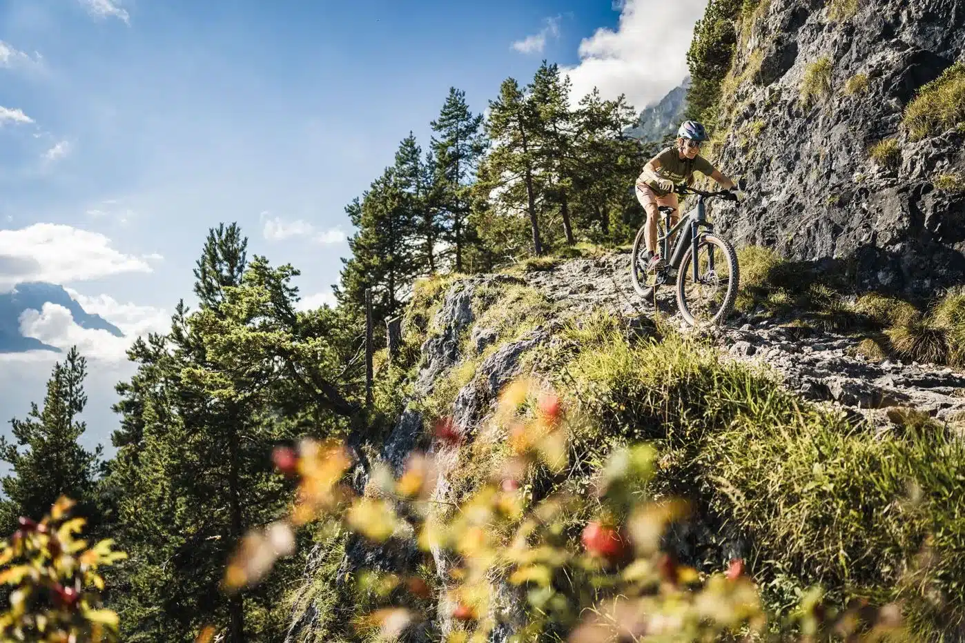 Eine Frau mit Helm und sportlicher Kleidung fährt mit einem E-Mountainbike auf einem felsigen Gebirgspfad. Im Hintergrund sind Bäume und eine Berglandschaft zu sehen.
