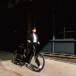 Elektrorad Stevens E-Comuna 7.4.1 HT, schwarz, mit Frau in Business-Kleidung und Helm, bereit für die Fahrt.