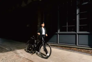 Elektrorad Stevens E-Comuna 7.4.1 HT, schwarz, mit Frau in Business-Kleidung und Helm, bereit für die Fahrt.