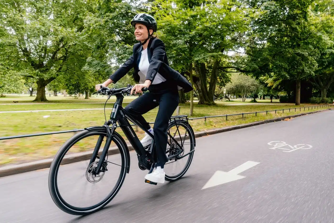Frau in schwarzem Anzug fährt ein Stevens E-Comuna 7.4.1 HT Fahrrad auf einem asphaltierten Weg im Park.