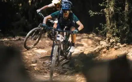 Eine sportliche Fahrerin mit Helm und Brille fährt mit einem modernen E-Mountainbike auf einem schmalen, steinigen Trail bergab, gefolgt von einer weiteren Person auf einem ähnlichen Rad.
