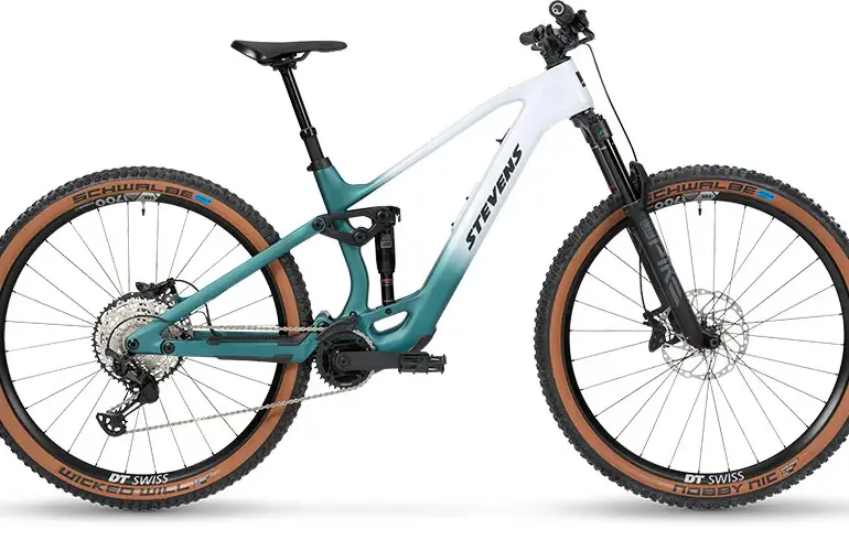 Seitenansicht des Stevens E-Maverick AM 7.4.3, einem weißen und türkisfarbenen E-Mountainbike mit Carbonrahmen und braunen Reifen.
