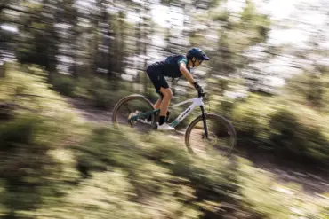 Mountainbiker fährt das weiße und grüne Stevens E-Maverick AM 7.4.3 bergab auf einem schmalen Waldweg, die Bäume und der Hintergrund sind durch die Bewegung unscharf.