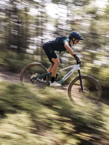 Mountainbiker fährt das weiße und grüne Stevens E-Maverick AM 7.4.3 bergab auf einem schmalen Waldweg, die Bäume und der Hintergrund sind durch die Bewegung unscharf.