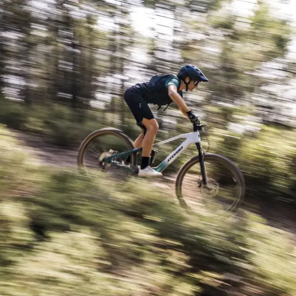Mountainbiker fährt das weiße und grüne Stevens E-Maverick AM 7.4.3 bergab auf einem schmalen Waldweg, die Bäume und der Hintergrund sind durch die Bewegung unscharf.