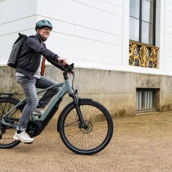 Mann mit Helm und Rucksack fährt ein Stevens E-Triton 7.8.1 Plus Forma E-Bike in River Stone Farbe vor einer Stuckfassade.