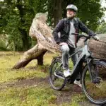 Mann mit Helm auf einem grauen Stevens E-Triton 7.8.1 Plus Forma E-Bike, sitzend auf einem großen, bemoosten Baumstamm im Freien, umgeben von Bäumen und Gras.
