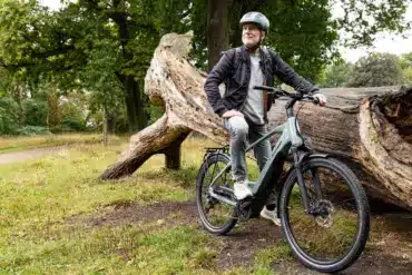 Mann mit Helm auf einem grauen Stevens E-Triton 7.8.1 Plus Forma E-Bike, sitzend auf einem großen, bemoosten Baumstamm im Freien, umgeben von Bäumen und Gras.