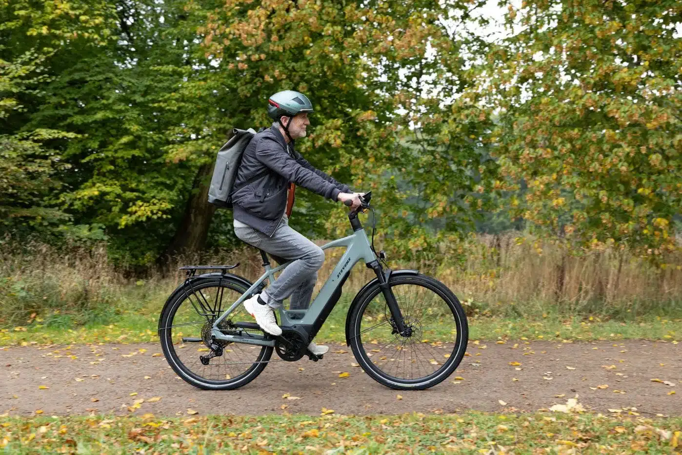 Ein Mann mit Helm und Rucksack fährt ein graues Stevens E-Triton 7.8.1 Plus Forma E-Bike auf einem Schotterweg im Wald.