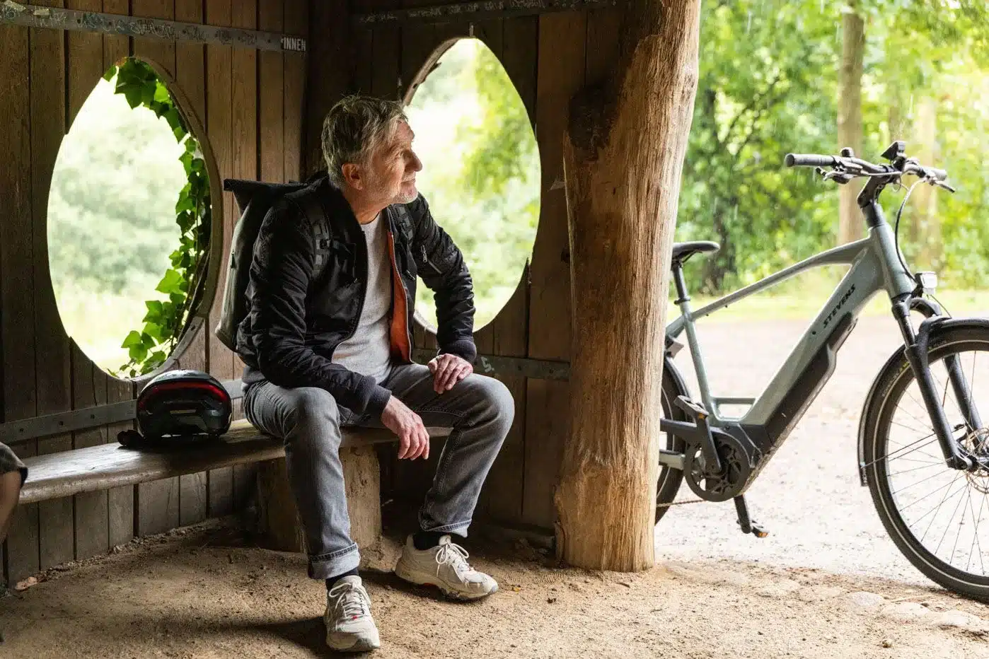 Mann mit Rucksack sitzt auf Holzbank und schaut nach rechts, daneben steht ein graues Stevens E-Triton 7.8.1 Plus Forma E-Bike.