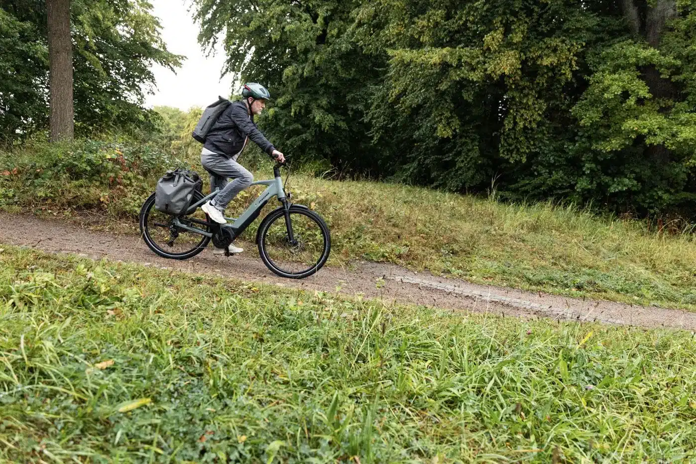 Ein Mann mit Helm und Rucksack fährt ein graues E-Bike auf einem Feldweg im Wald.