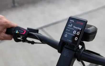Das Bosch Kiox 500 Display eines E-Bikes zeigt Fahrmodi wie 'Turbo' und 'Touring', Akkustand und Bremsweg. Links daneben ist die Bedieneinheit mit verschiedenen Tasten und Anzeigen zu sehen.