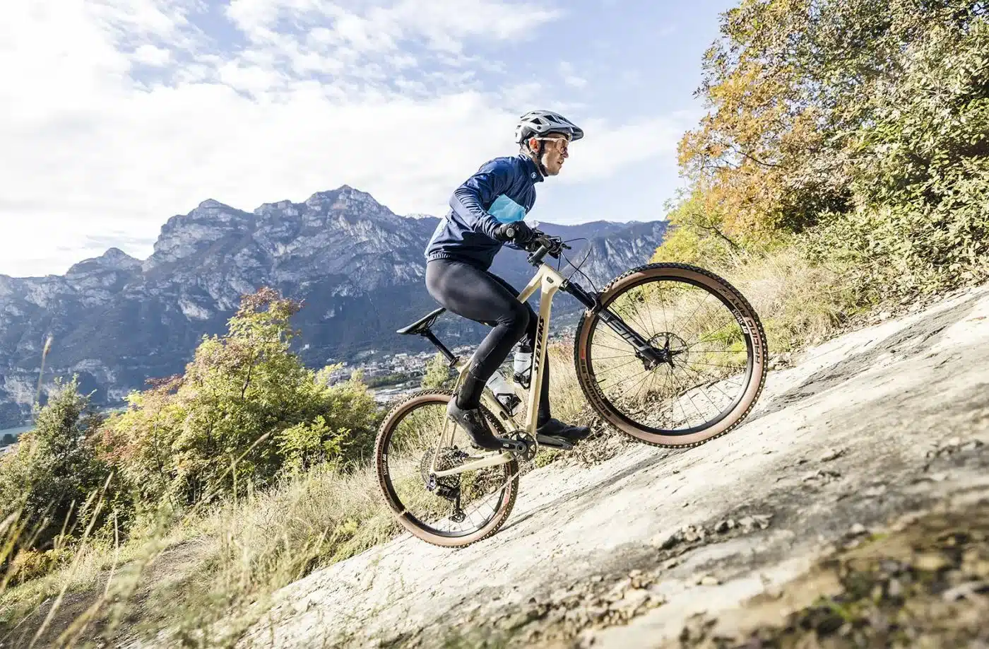 Fahrer in blauer Jacke und schwarzer Hose fährt ein sandfarbenes Mountainbike auf einem felsigen Hang mit Bergpanorama im Hintergrund.