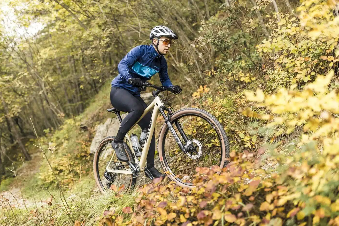Eine Frau mit Helm und Sportbekleidung fährt ein hellbraunes Stevens Sonora GX AXS Mountainbike einen mit Laub bedeckten Waldweg bergab.