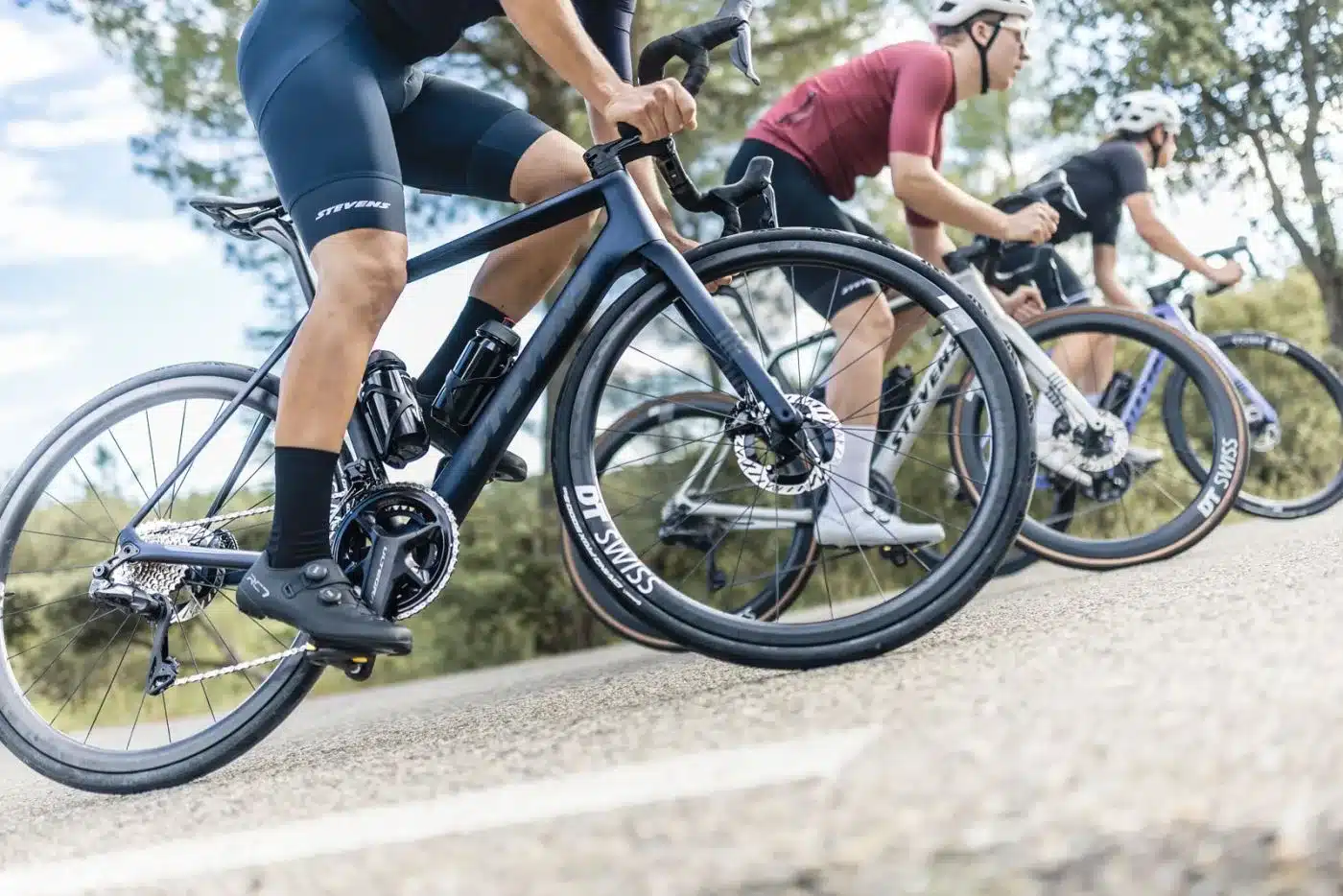 Drei Radfahrer fahren nebeneinander auf der Straße, wobei das vordere Stevens Xenith Rennrad im Fokus steht. Das Rad ist dunkelgrau und hat eine schlanke Silhouette.