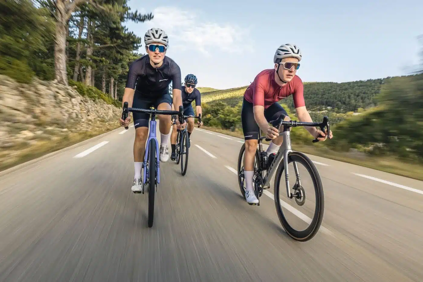 Drei Personen fahren nebeneinander auf Rennrädern auf einer asphaltierten Straße mit Bäumen und Hügeln im Hintergrund.