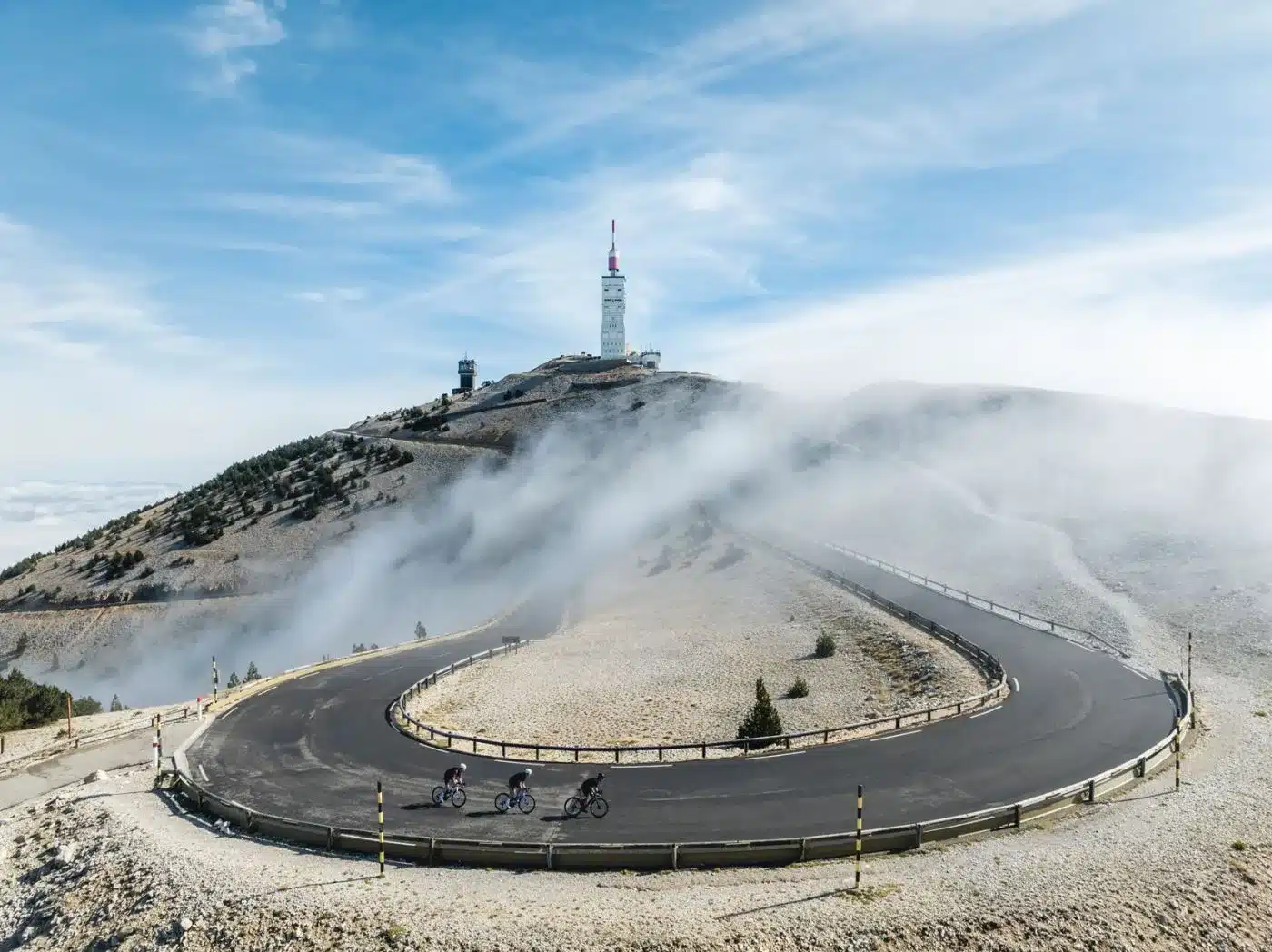 Drei Rennradfahrer auf einer Serpentinenstraße in den Bergen, mit einem Sendeturm in der Ferne und Nebelschwaden.