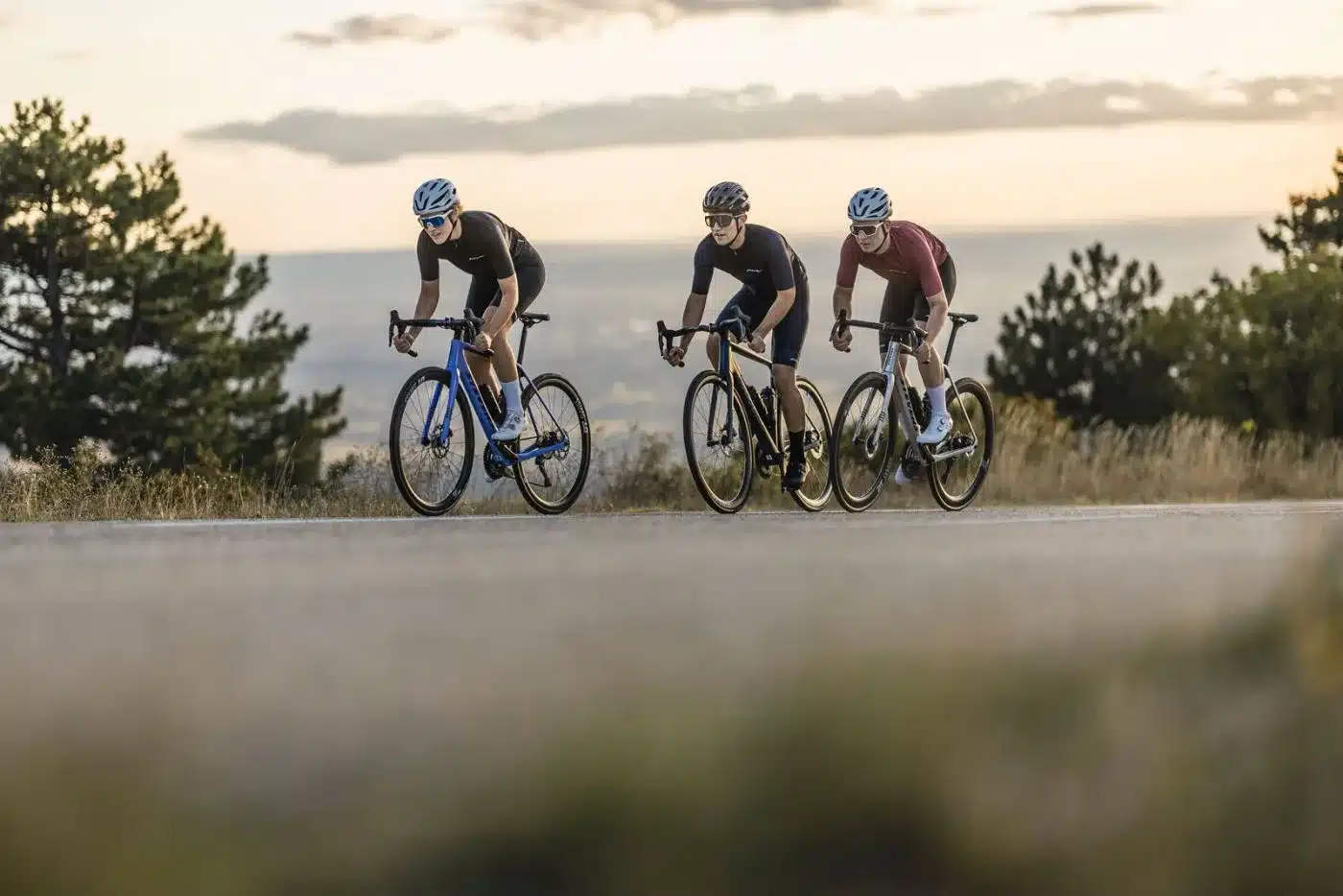 Drei Radfahrer in sportlicher Kleidung fahren auf der Straße, die Landschaft im Hintergrund ist leicht unscharf.