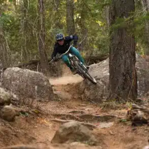 Mountainbikerin fährt auf einem staubigen Trail bergab, umgeben von Bäumen und Felsen.