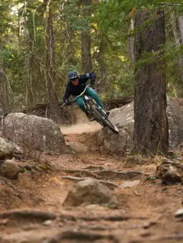 Mountainbikerin fährt auf einem staubigen Trail bergab, umgeben von Bäumen und Felsen.
