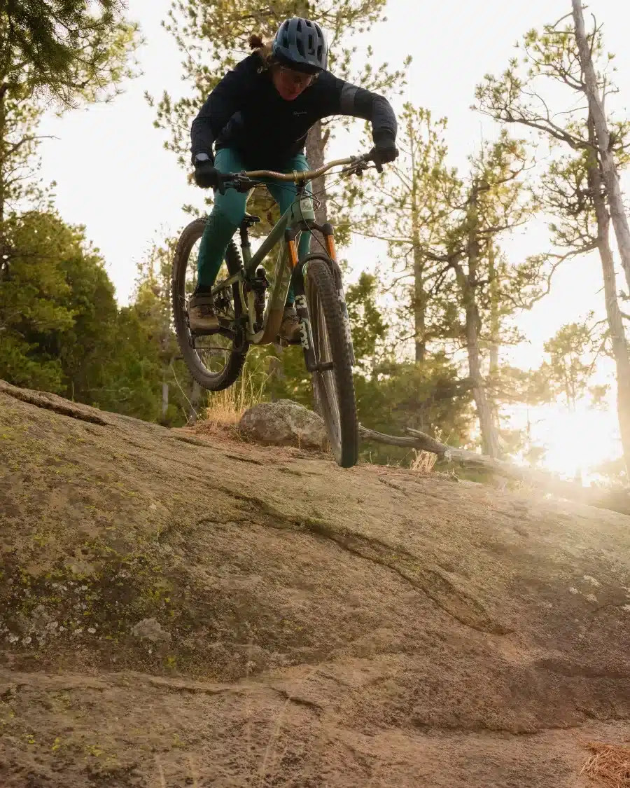Eine Mountainbikerin mit Helm und sportlicher Kleidung fährt mit einem Vollfederrahmen-Mountainbike über einen felsigen Abhang im Wald.