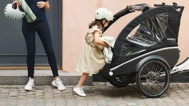 Kind mit Helm steigt in die Kabine eines schwarzen Kangaroo Lite Dreirad-Lastenrads, daneben steht eine Person mit weiterem Helm.